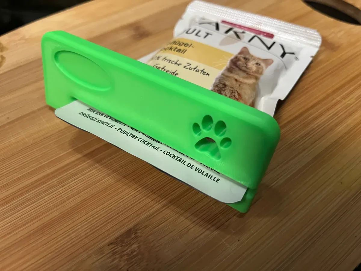 The green squeezer with a paw print sits on a cat food pouch, the pouch lying on a wooden board