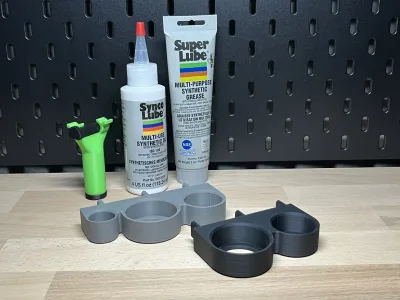 Zwei graue Halter auf der Holzplatte vor der schwarzen Lochwand, dahinter die Flaschen Super Lube und Synco Lube sowie eine grüne Fettpresse