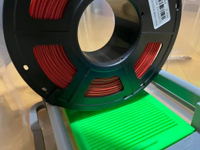 Eine schwarze Spule mit rotem Filament steht im Halter, darunter die grüne Box mit ihrem gerippten Deckel