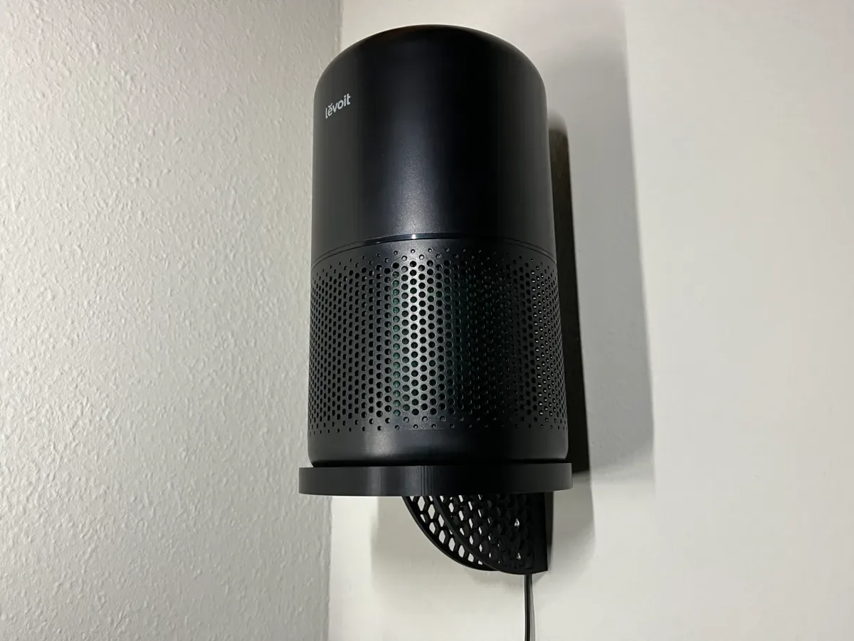 The black air purifier stands on the round wall shelf against a white wall, the cable leaving the channel at the bottom